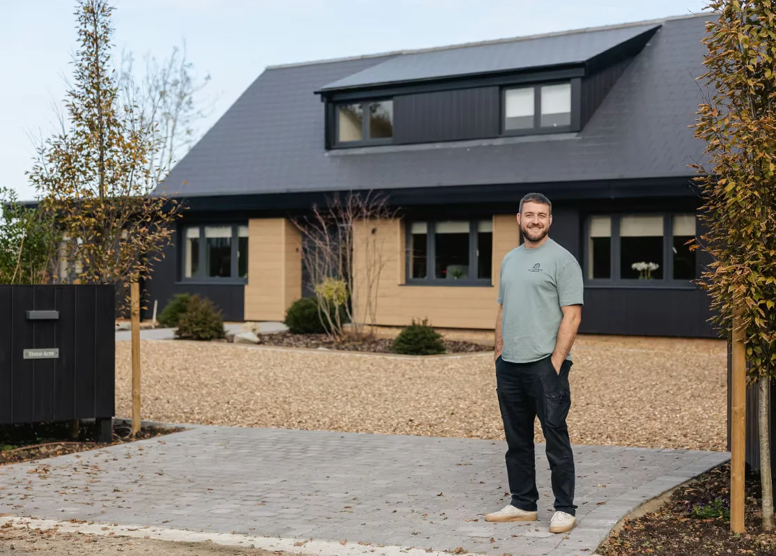 A man from ALC Carpentry and Construction in East Suffolk standing in front of a modern and large house in a driveway smiling.